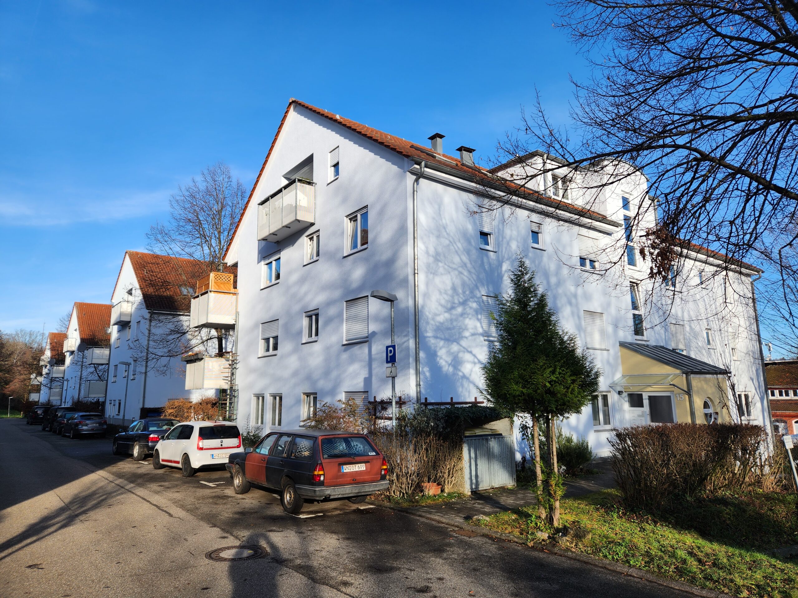 Mehrfamilienhaus mit weißen Fassaden und roten Dächern an einer Wohnstraße, geparkte Autos und winterliche Vegetation bei blauem Himmel.