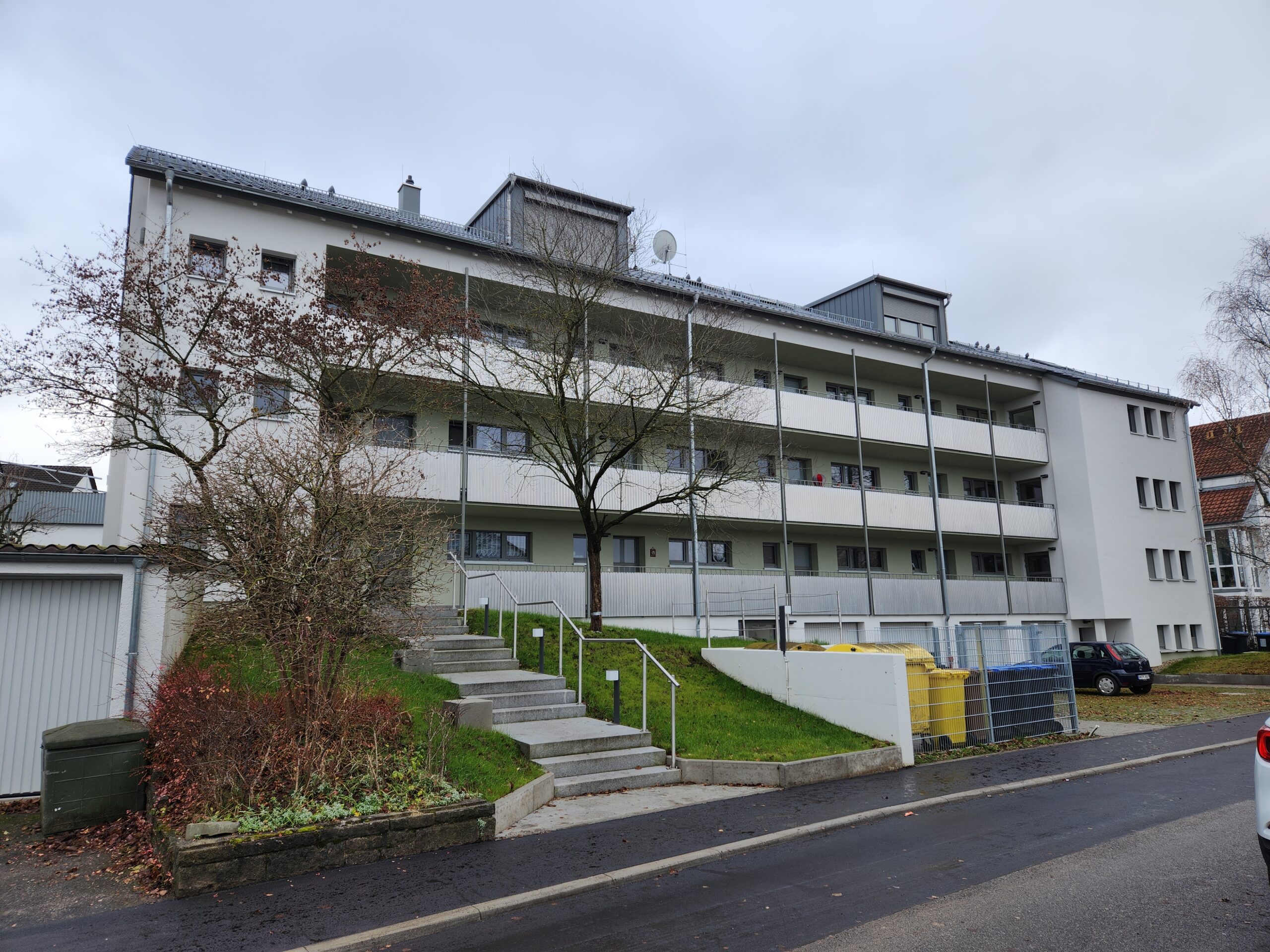 Mehrstöckiges Wohngebäude mit Balkonen und hellgrauer Fassade, umgeben von Bäumen und geparkten Autos.