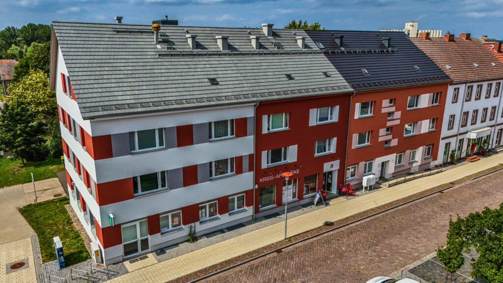 Modernisiertes Wohn- und Geschäftsgebäude mit rot-weißer Fassade, grauem Dach und einer Apotheke im Erdgeschoss an einer gepflasterten Straße.