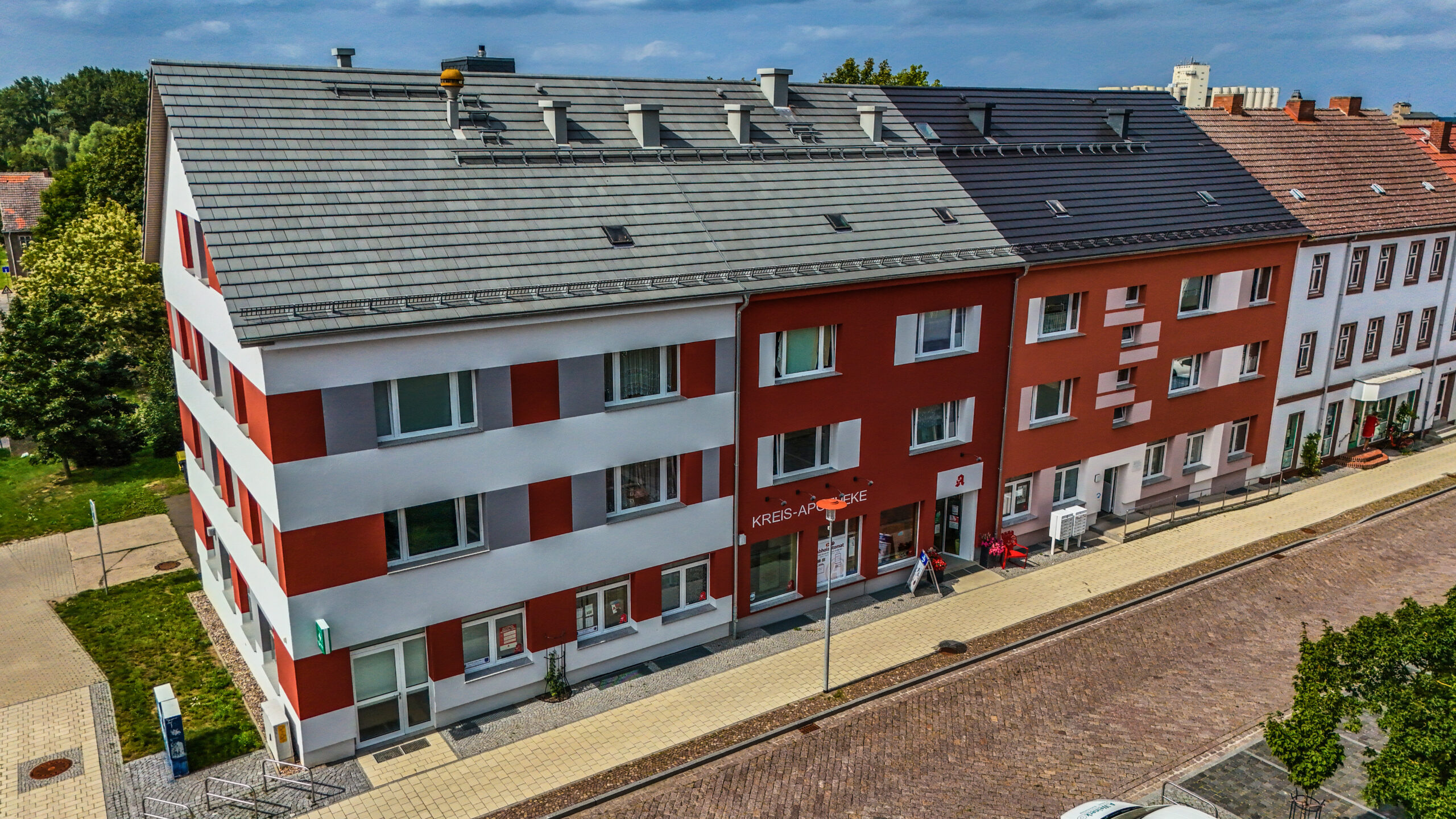 Modernisiertes Wohn- und Geschäftsgebäude mit rot-weißer Fassade, grauem Dach und einer Apotheke im Erdgeschoss an einer gepflasterten Straße.