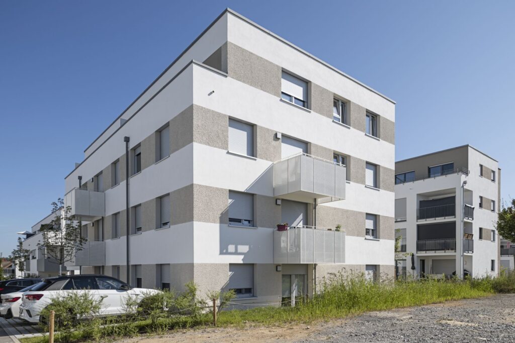 Modernes Mehrfamilienhaus mit klarer Linienarchitektur, heller Fassade und Balkonen, daneben geparkte Autos unter blauem Himmel.